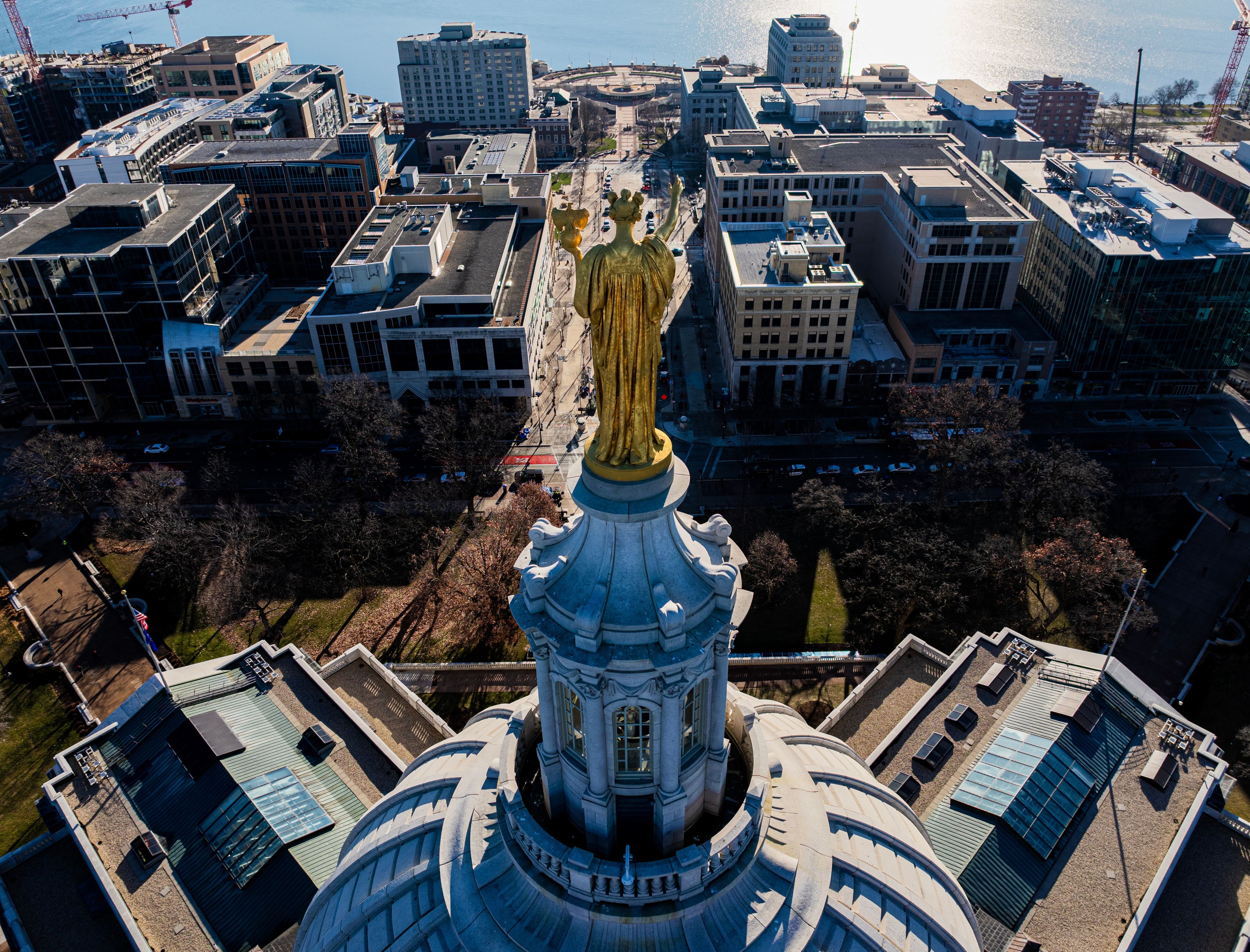 Aerial drone view looking down past the golden Wisconsin statue atop the dome of the Wisconsin State Capitol building toward downtown Madison. High-angle travel and landmark photography.
