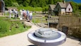 Un sitio histórico con un modelo de noria, una casa de madera y un grupo de personas observando las exhibiciones.