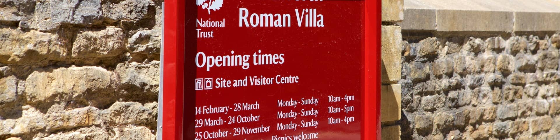 Un letrero rojo con texto blanco para la Villa Romana de Chedworth, que incluye los horarios de apertura y un mensaje de bienvenida.