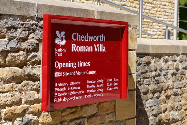 Ein rotes Schild mit weißer Schrift für die Chedworth Roman Villa, inklusive Öffnungszeiten und einer Willkommensbotschaft.