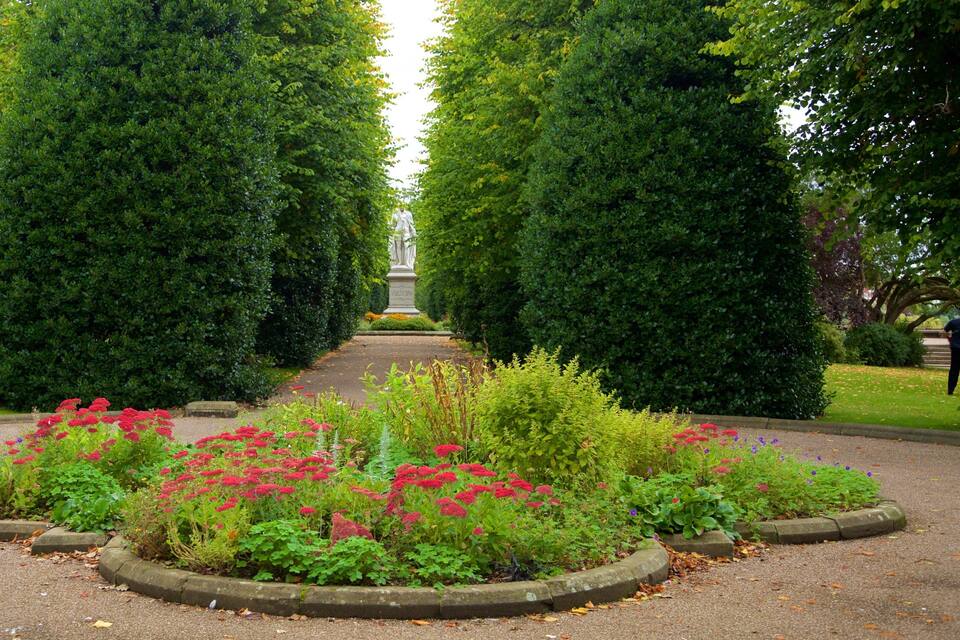 A formal garden with a central statue, surrounded by neatly trimmed hedges and vibrant flower beds.
