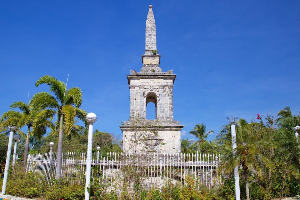 A historical monument with a tall obelisk, surrounded by a white fence and palm trees.