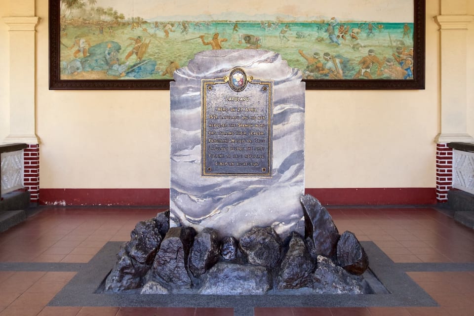 A historical monument with a plaque, surrounded by a stone base and a mural depicting a battle scene.