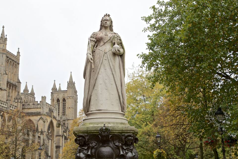 A statue of a queen on a pedestal in a park with a historic building in the background.