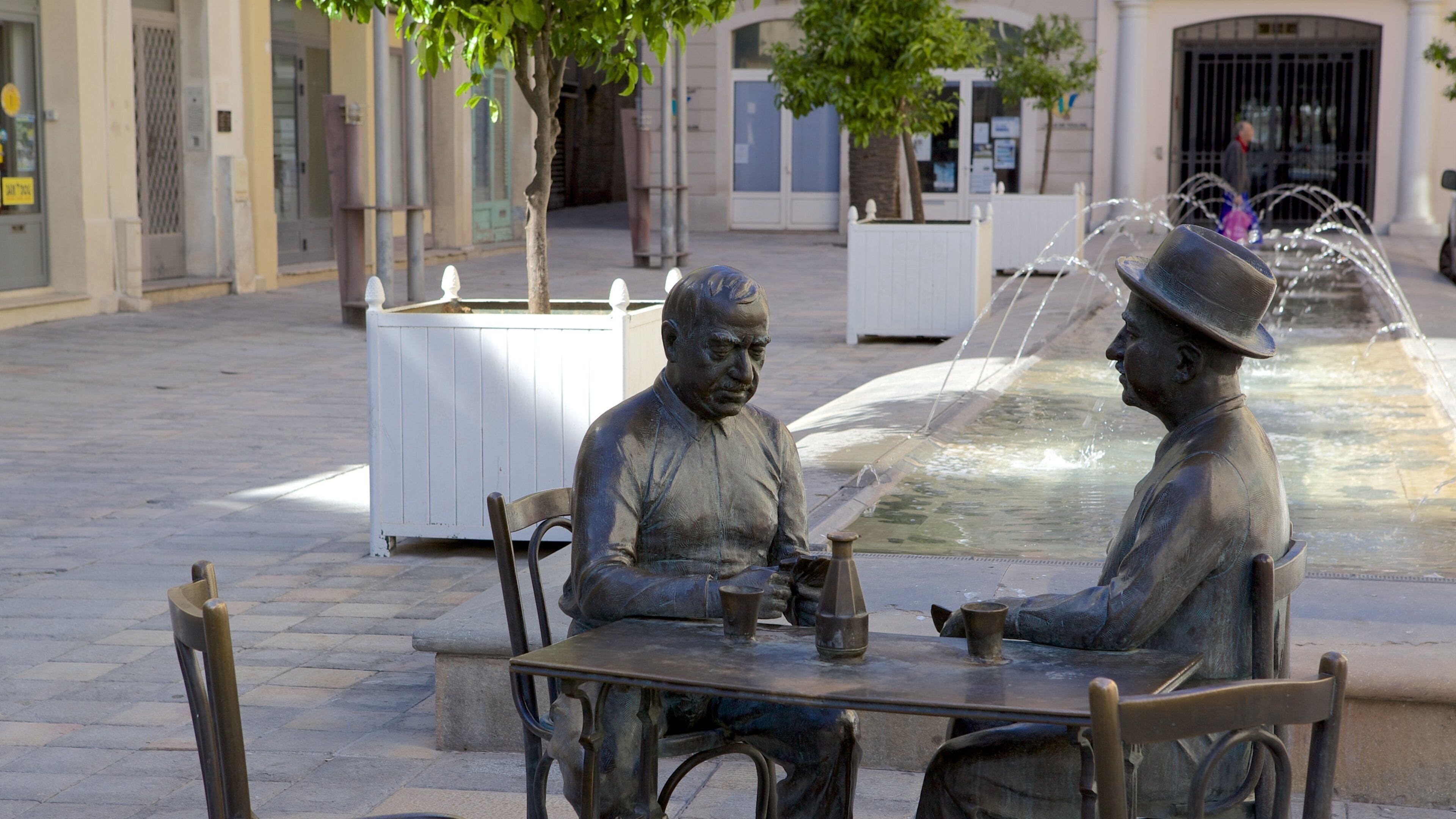 A statue of two men seated at a table with a fountain in the background.