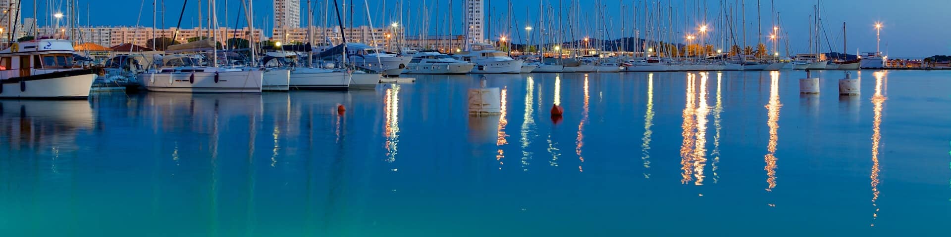 A marina at dusk with numerous sailboats docked, reflecting city lights on the water.