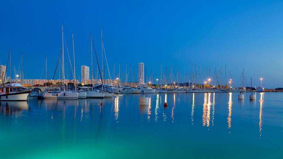 A marina at dusk with numerous sailboats docked, reflecting city lights on the water.