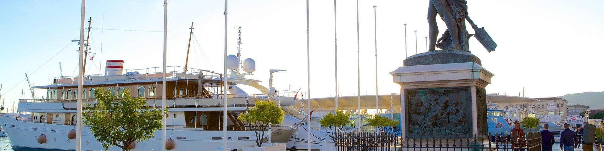 Una estatua de un hombre señalando, rodeada por una cerca, con barcos amarrados al fondo.