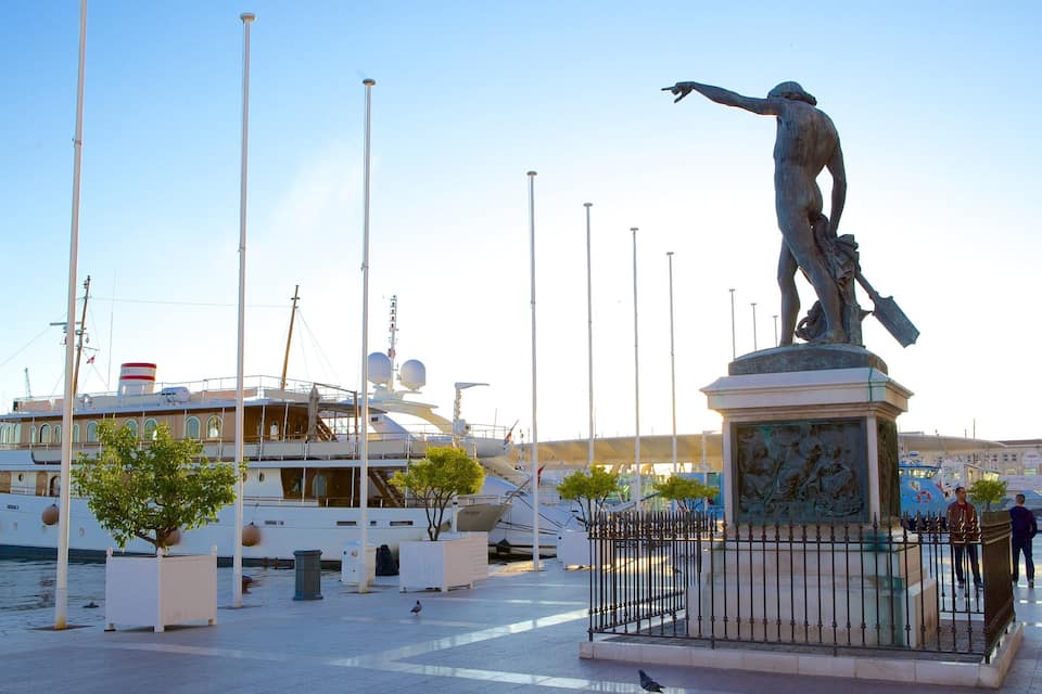 Une statue d’un homme pointant du doigt, entourée d’une clôture, avec des bateaux amarrés en arrière-plan.