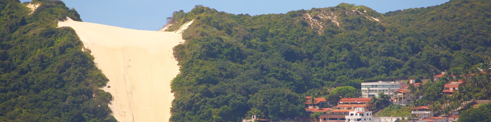 Paisaje costero con una duna de arena destacada, colinas verdes exuberantes y una zona de playa con edificios y sombrillas.
