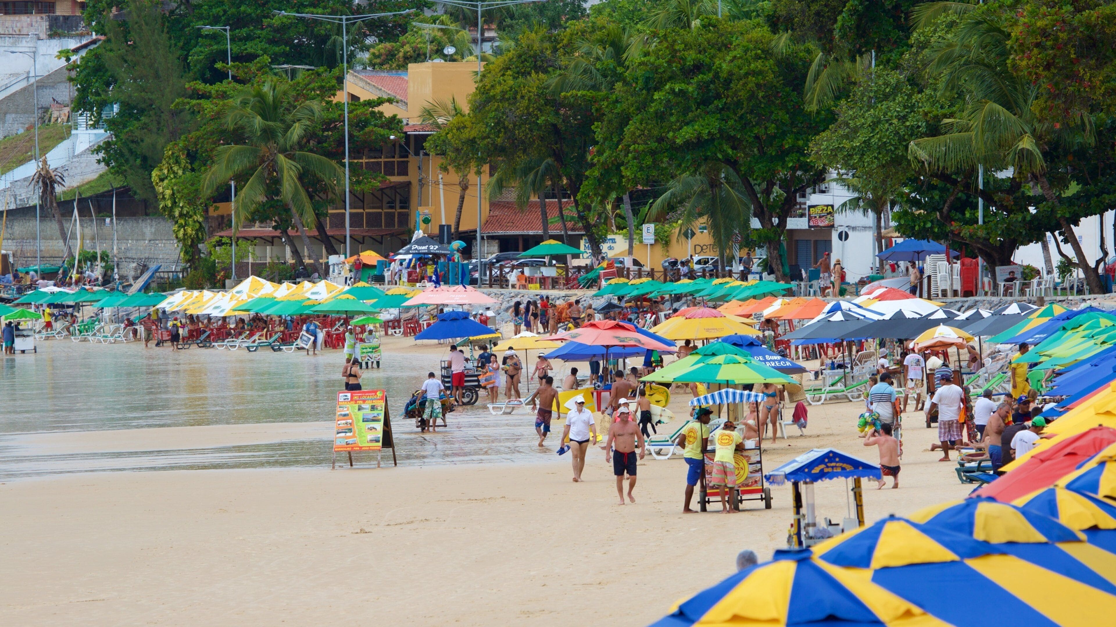 Une plage bondée, avec des parasols aux couleurs vives et des gens qui profitent du sable et de l’eau.