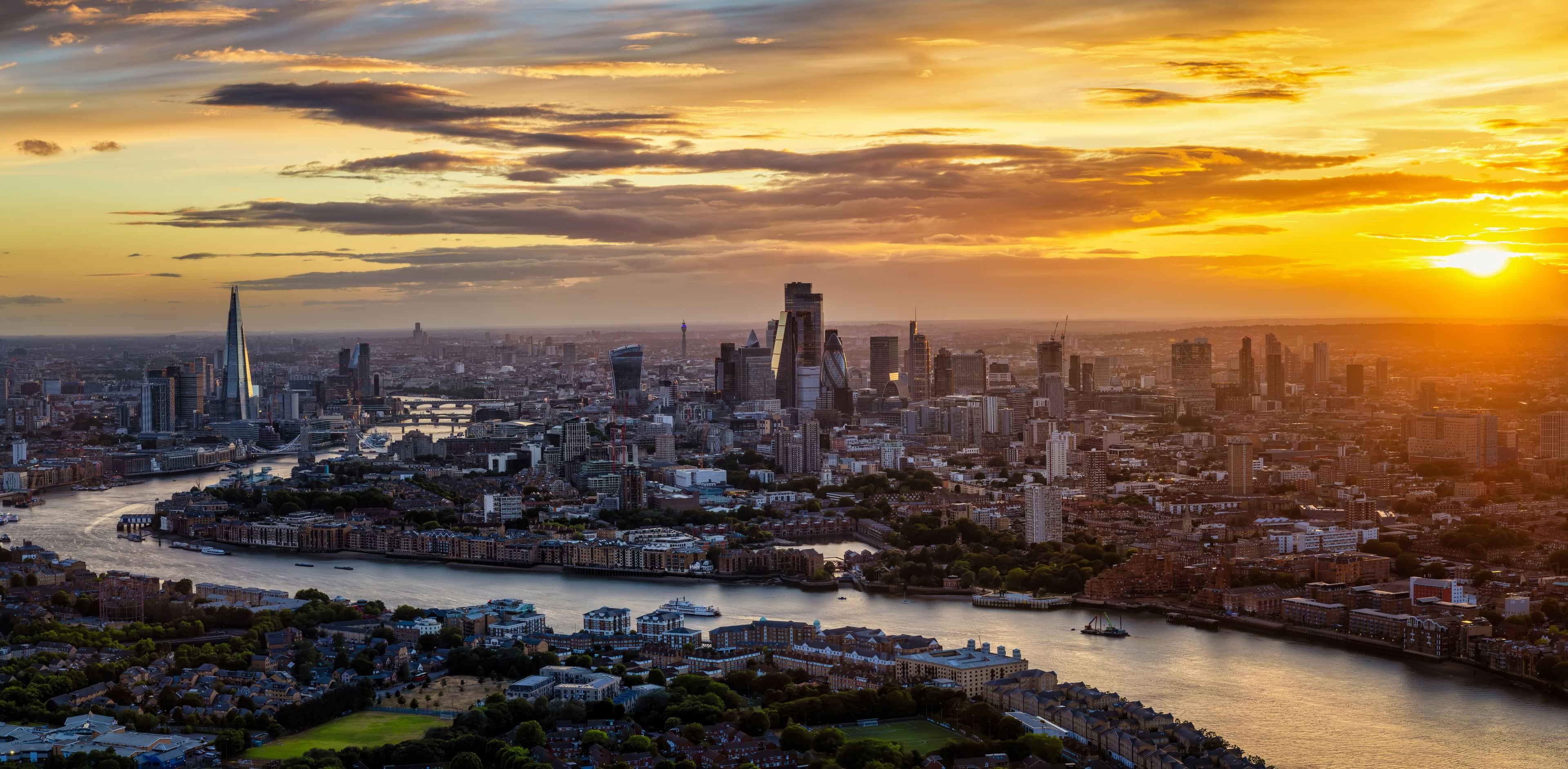 Une vue sur une ville au coucher du soleil, avec une rivière qui la traverse, mettant en valeur des gratte-ciel modernes et des bâtiments historiques.