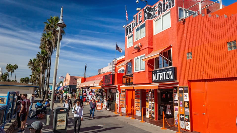 A vibrant street with a red building labeled "MUSELE BEACH NUTRITION" and palm trees in the background.