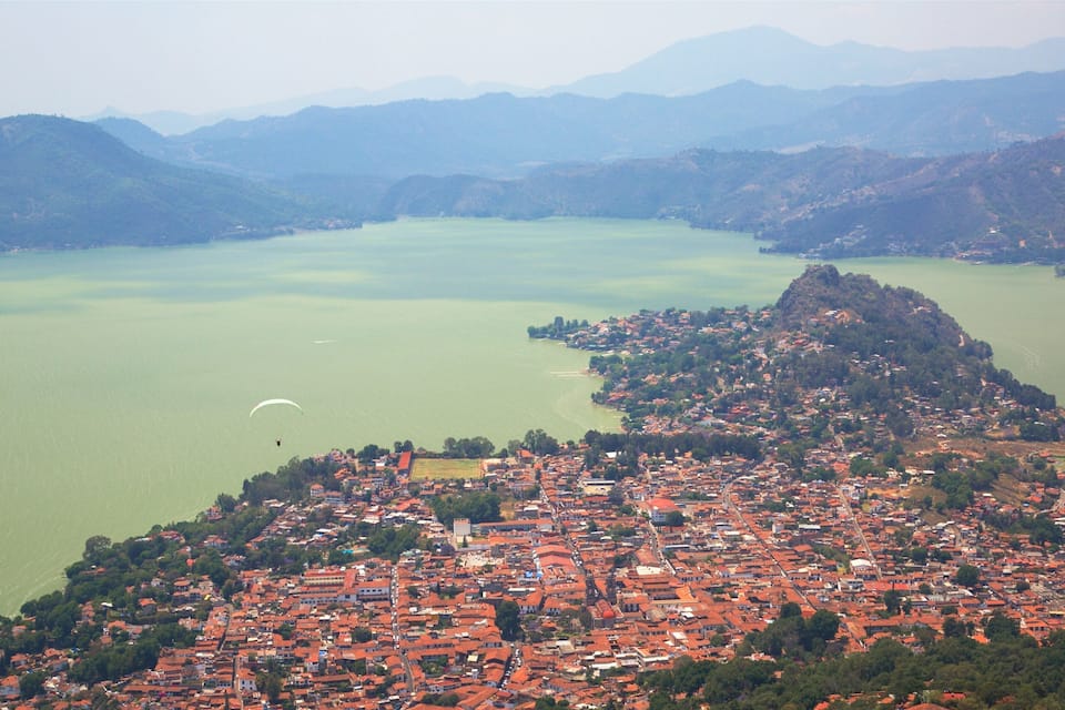 Un pueblo costero con edificios de techo rojo, un lago verde y montañas al fondo.