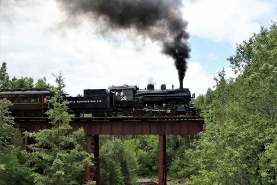 A steam locomotive on a bridge emitting black smoke, surrounded by dense greenery.