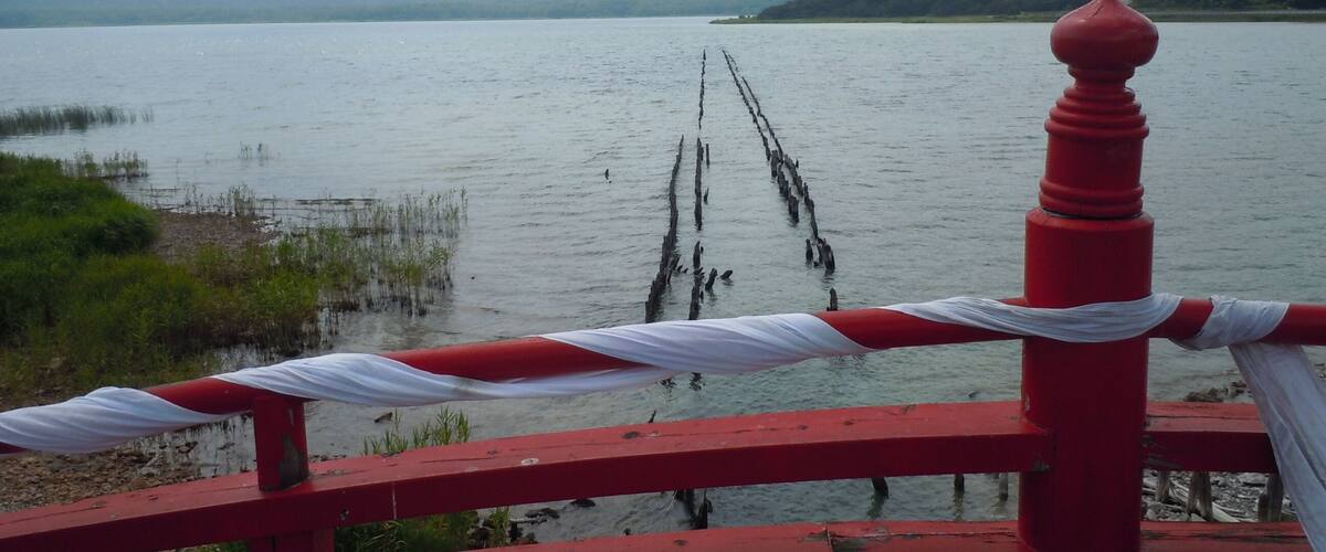A wooden bridge with red railings and white drapes, leading over a calm lake with visible wooden stakes in the water.