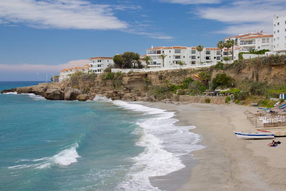 A coastal beach with clear turquoise waters, a sandy shore, and a row of white buildings with red roofs on a cliff.