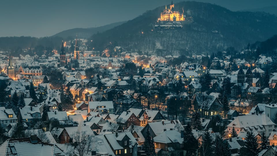 A snow-covered town with a prominent castle on a hill, surrounded by residential houses and trees.