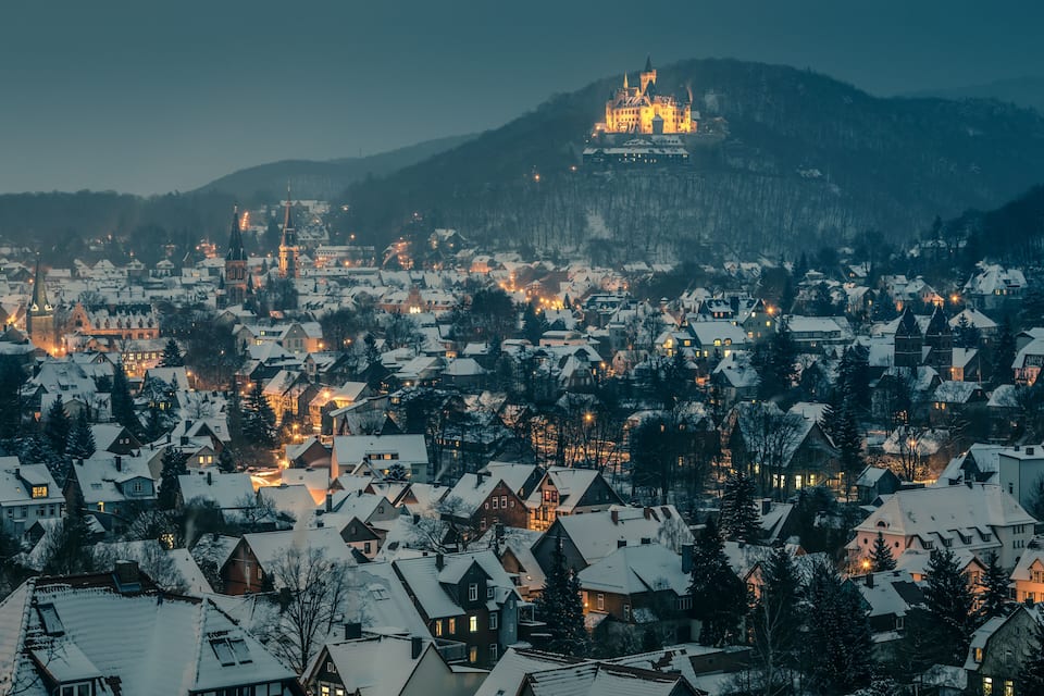 A snow-covered town with a prominent castle on a hill, surrounded by residential houses and trees.