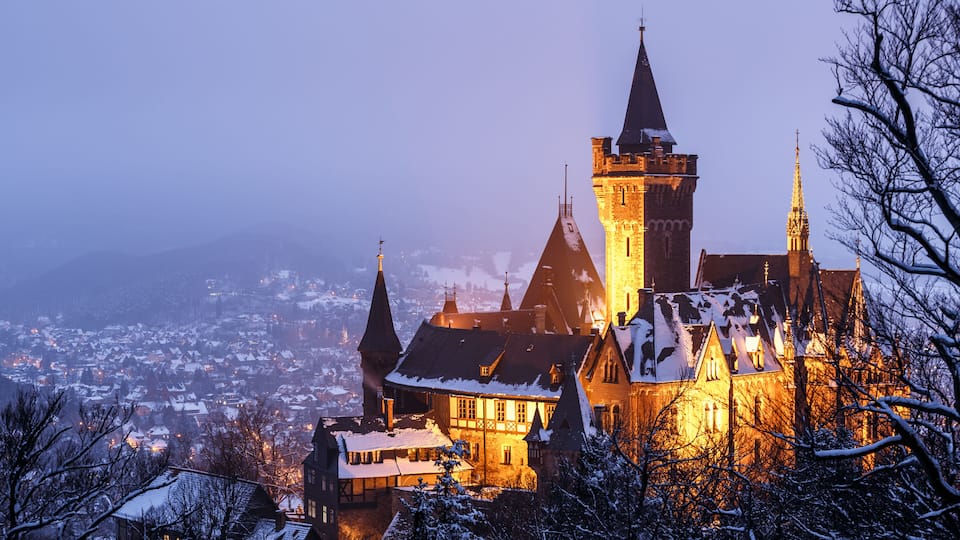 A historic castle with multiple towers, surrounded by a snowy landscape and a town in the background.