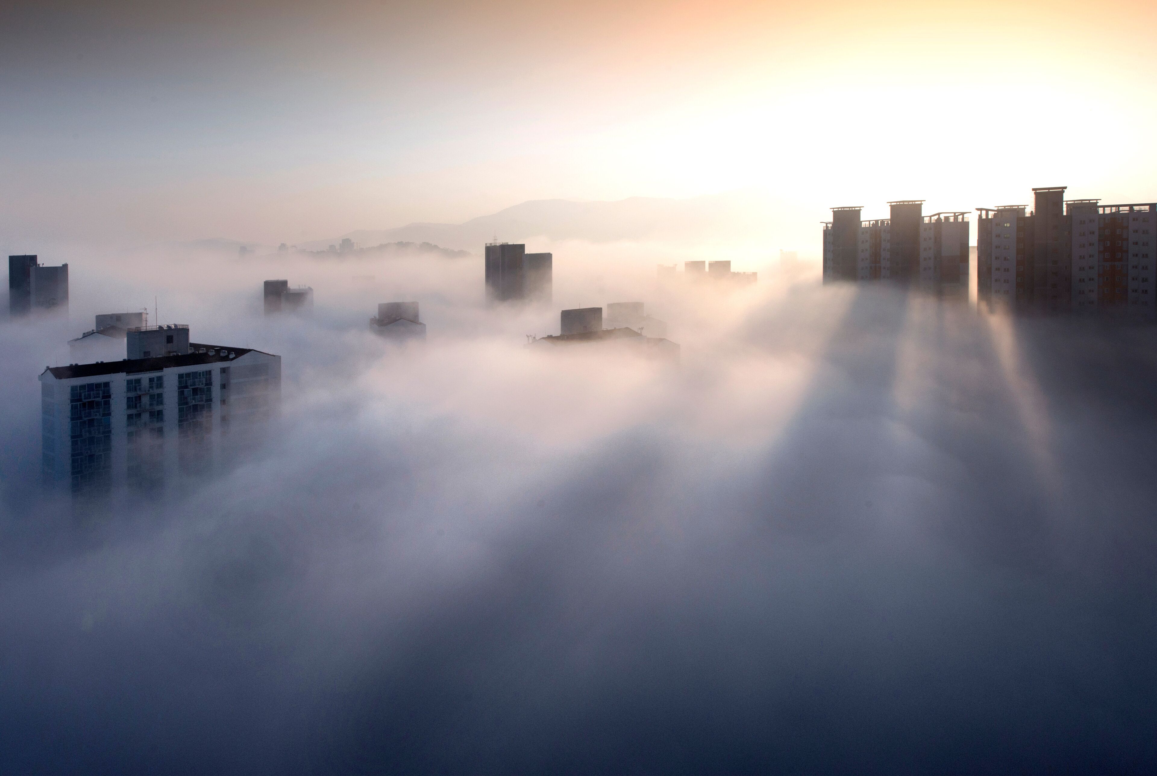 A cityscape with buildings partially obscured by fog, with sunlight filtering through.
