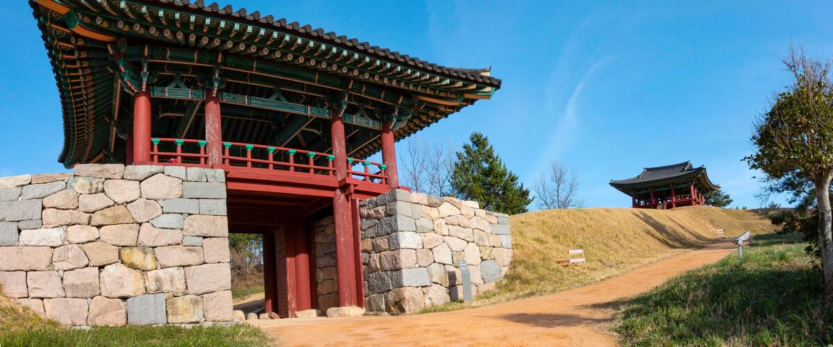 A traditional Korean gate with a stone base and red pillars, leading to a temple on a hill.