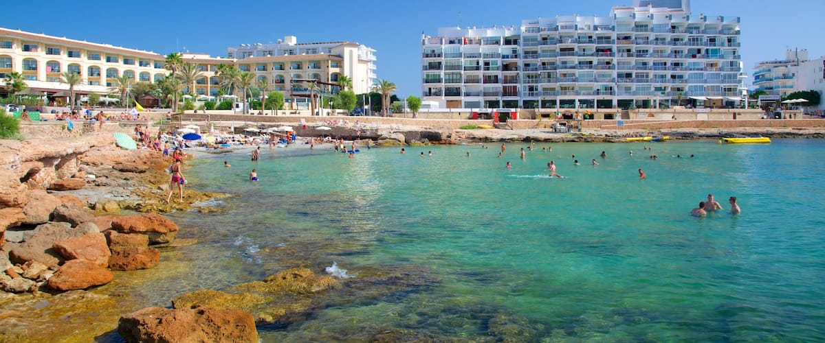 A beach with clear turquoise water, rocky shore, and modern buildings in the background.