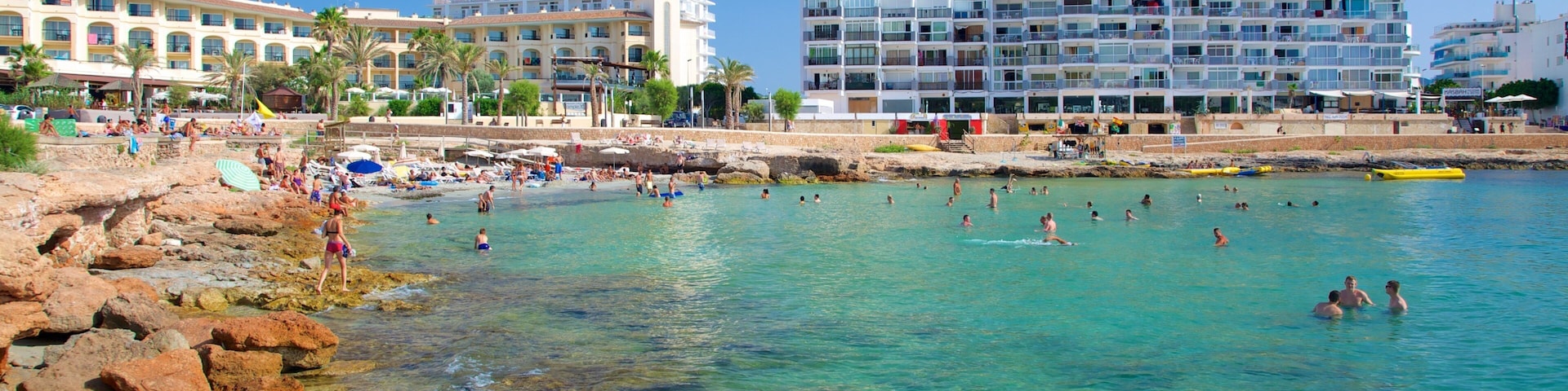 A beach with clear turquoise water, rocky shore, and modern buildings in the background.