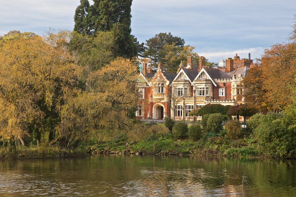 A large, ornate Victorian-style mansion surrounded by dense trees and a calm lake.