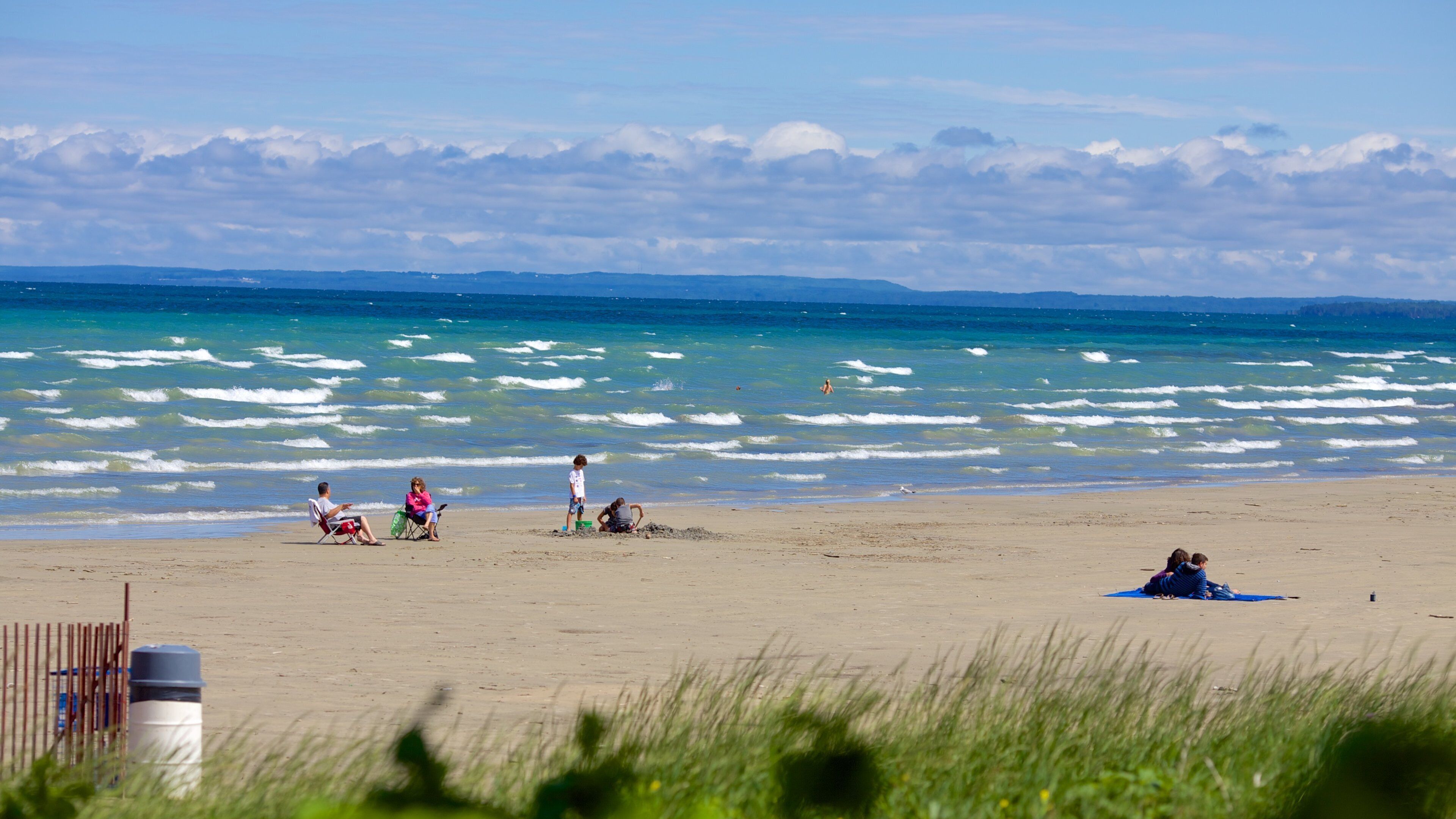 A beach with people relaxing and playing in the sand, waves crashing onto the shore, and a clear sky.