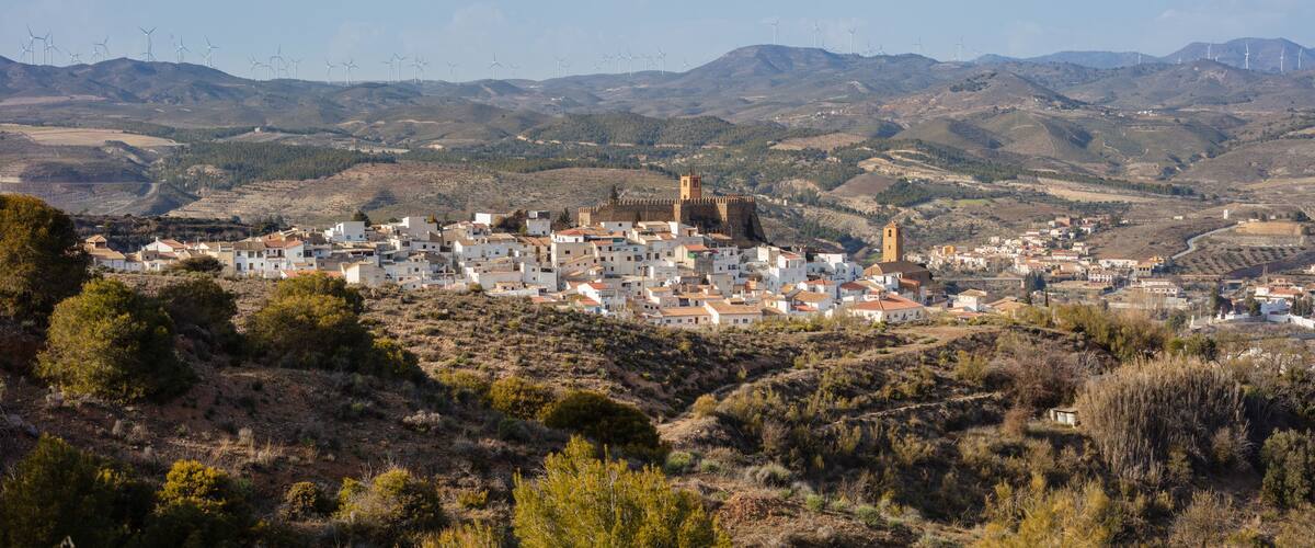 A coastal town with white buildings, a prominent tower, and wind turbines in the background.
