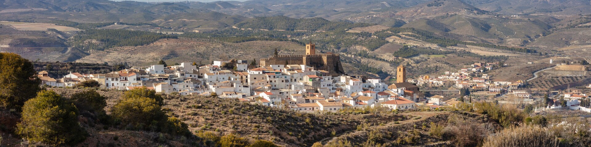 A coastal town with white buildings, a prominent tower, and wind turbines in the background.