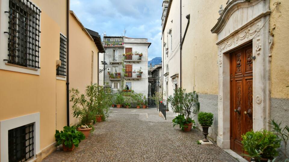 A cobblestone street flanked by buildings with balconies and potted plants.