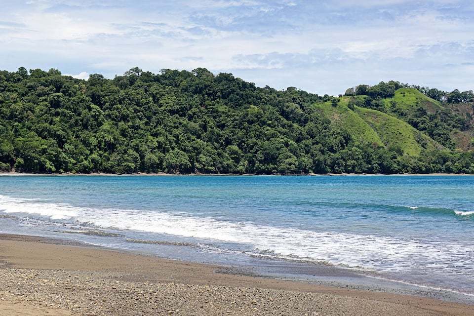 A beach with waves, a forested hill, and boats in the water.