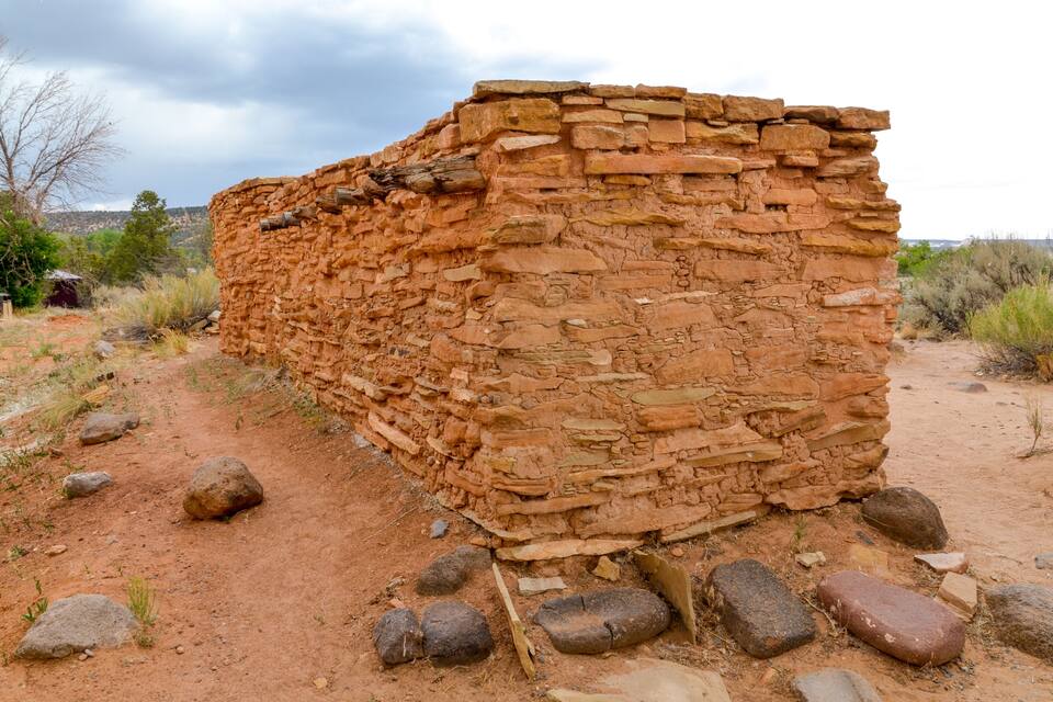 Une vaste structure de pierre ancienne, à l’aspect rugueux et aux couches apparentes, entourée d’un paysage désertique où la végétation est rare et où l’on trouve des rochers éparpillés.