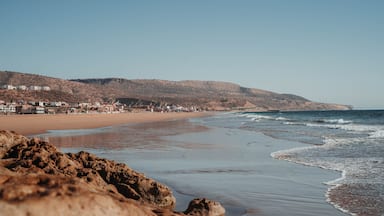 A beach with rocky foreground, sandy shore, and buildings in the background.