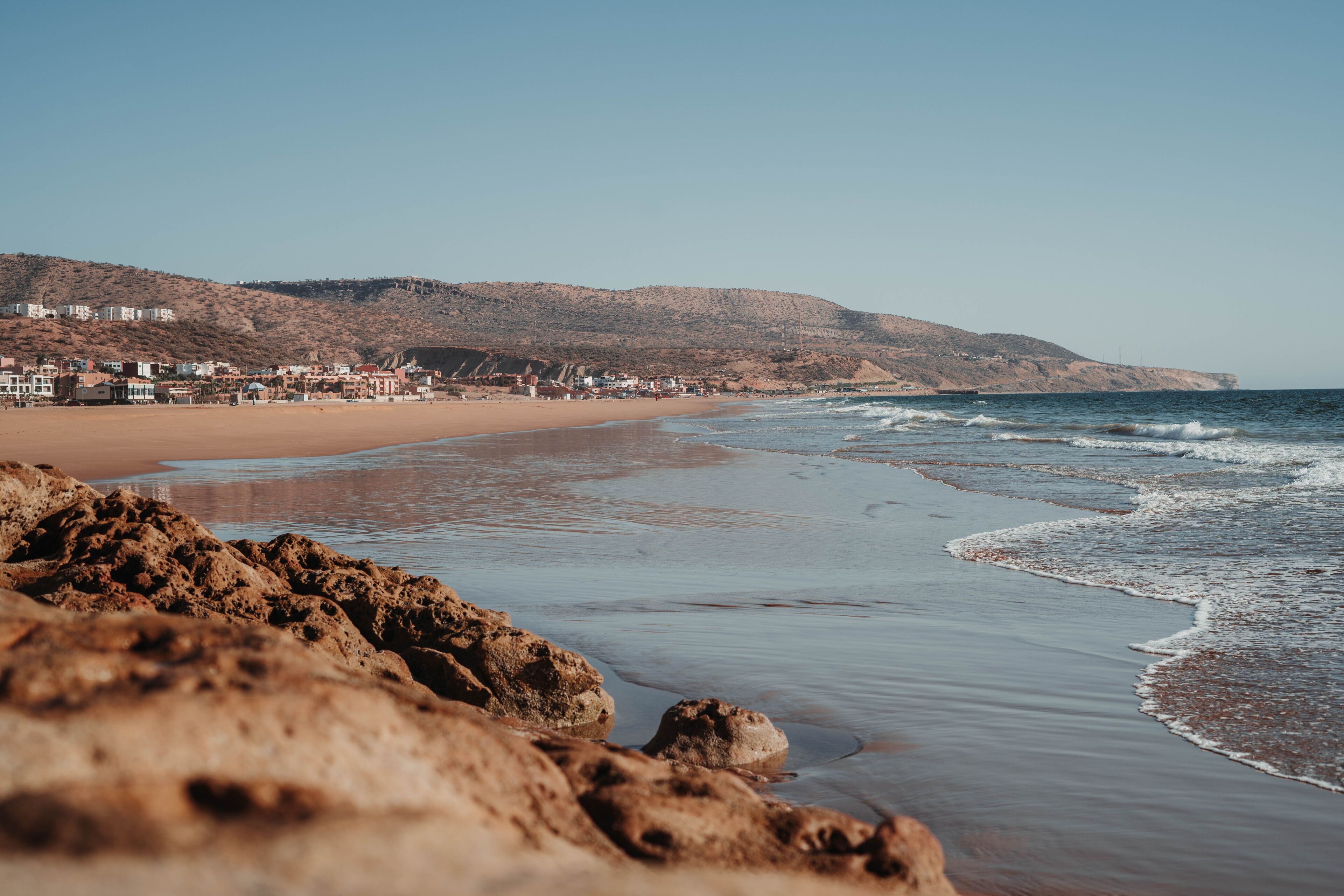 A beach with rocky foreground, sandy shore, and buildings in the background.