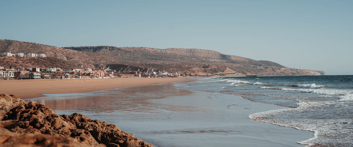 A beach with rocky foreground, sandy shore, and buildings in the background.