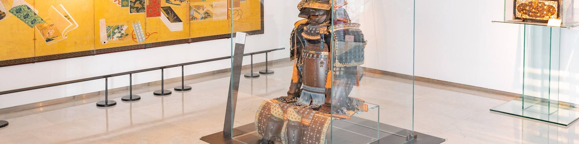 A museum exhibit with a glass display case containing a samurai armor and a large painting on the wall.