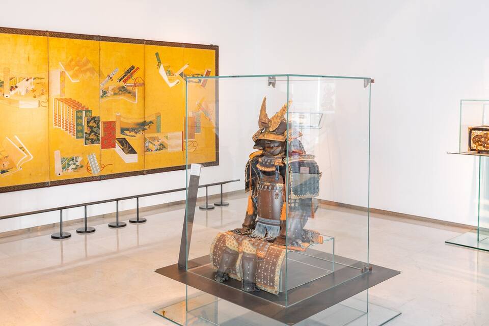 Uma exposição de museu com uma vitrine de vidro que contém uma armadura samurai e uma grande pintura na parede.
