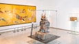 Una exhibición de museo con una vitrina de vidrio que contiene una armadura de samurai y una pintura grande en la pared.