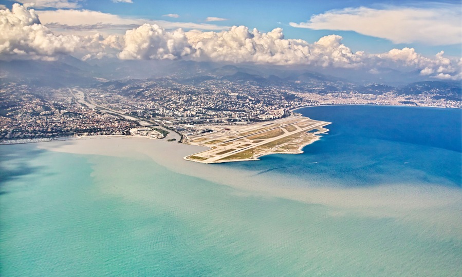 Vista aerea di una città costiera con un grande aeroporto, acque blu limpide e montagne sullo sfondo.
