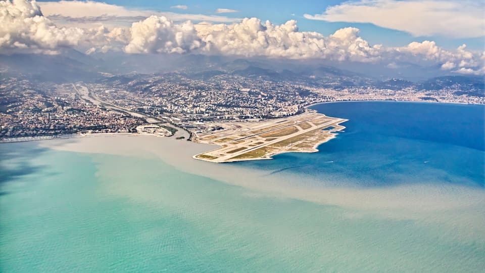 Vista aérea de uma cidade costeira com um grande aeroporto, águas azuis cristalinas e montanhas ao fundo.