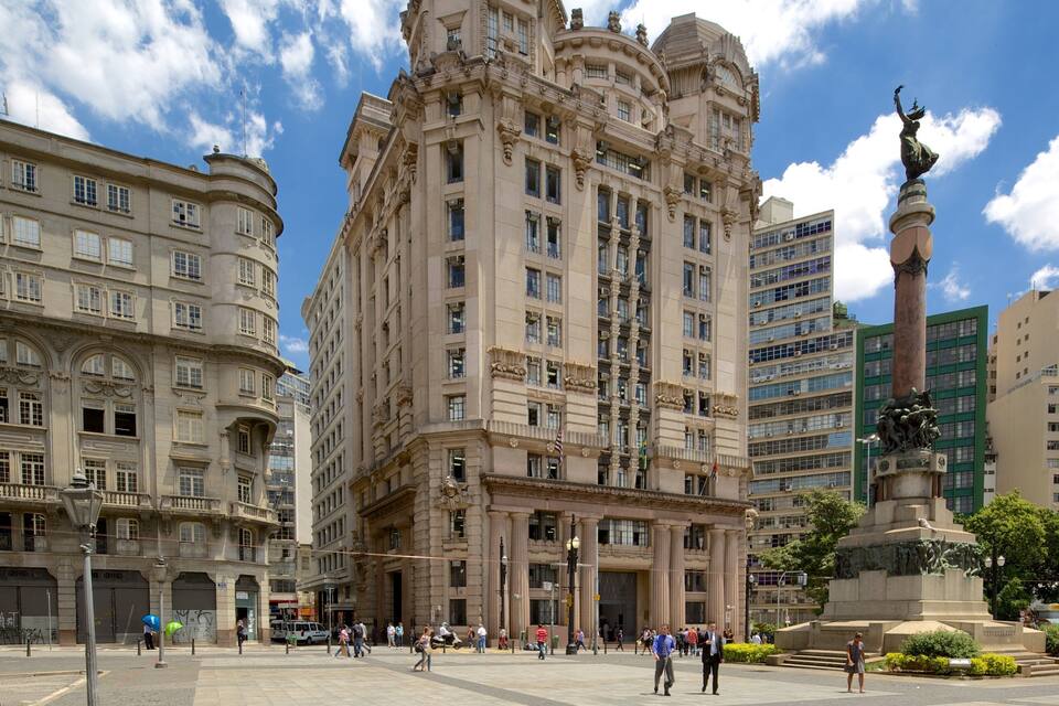 A historic building with ornate architecture, a statue, and a plaza with pedestrians.
