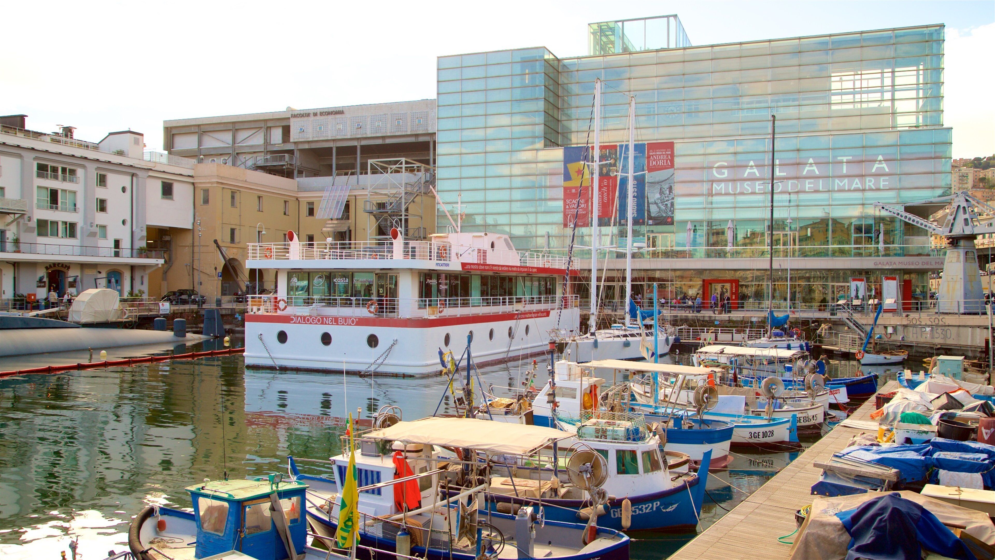 A marina with various boats docked, a large glass building labeled "Galata MUSEO DEL MARE," and a building with "FACULTY OF ECONOMICS" signage.