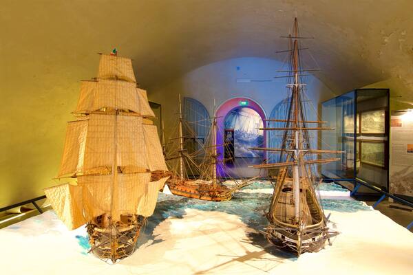 Deux grands voiliers sont exposés dans un musée, avec en arrière-plan une roue de navire et une fresque murale sur le thème maritime.