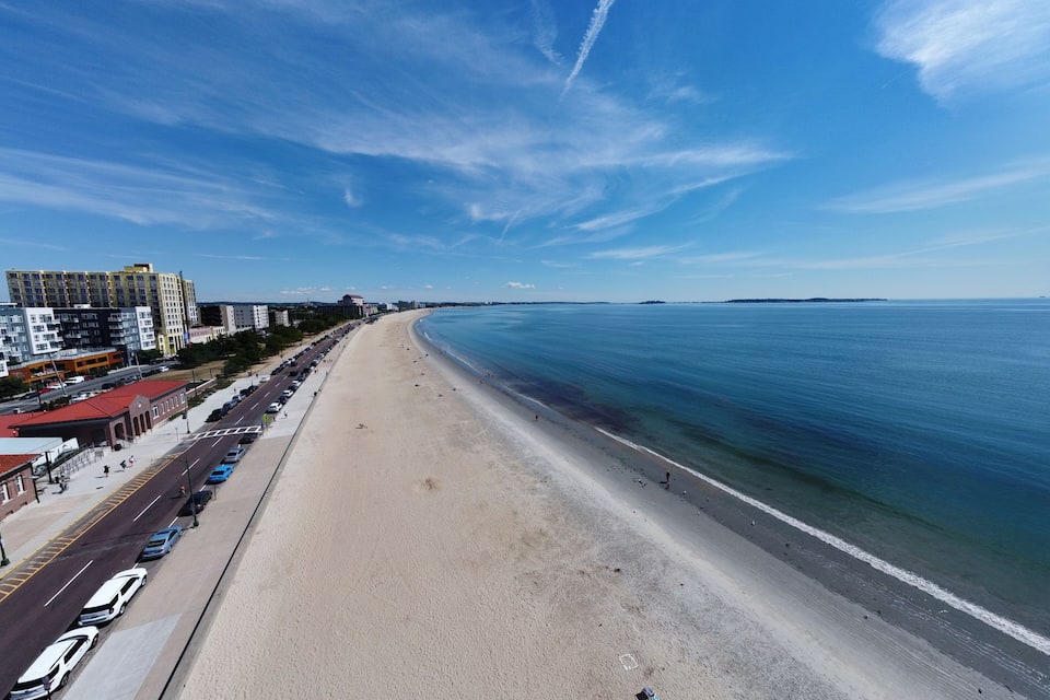A coastal cityscape with a wide sandy beach, a clear blue sky, and a distant island.