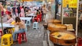 A bustling street market with people dining at tables, food stalls, and large cooking pots.