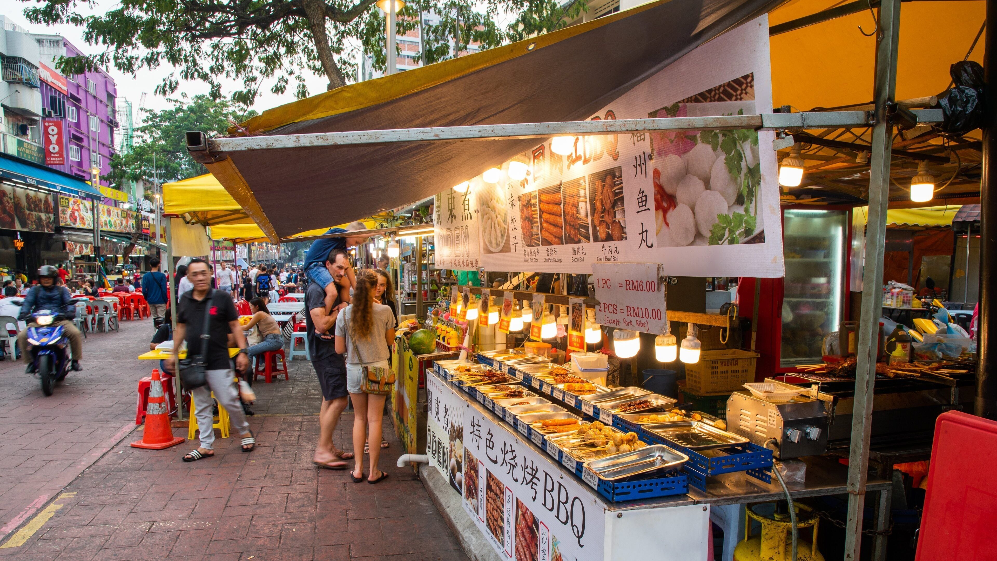 Un marché animé où l’on trouve de nombreux stands de nourriture de rue, des gens qui mangent sur place et des motos qui passent.