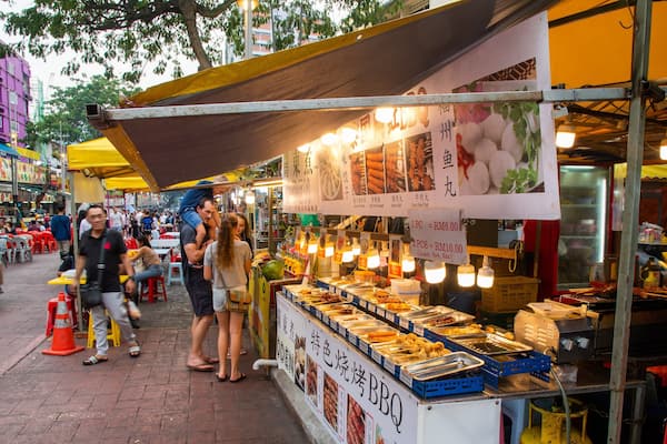 Mercado de comida de rua movimentado, com diversos pontos de venda de alimentos, pessoas almoçando e motocicletas passando.